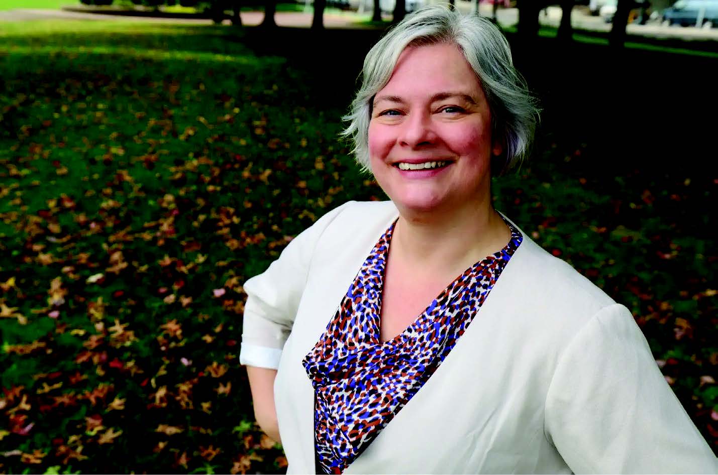 Picture of PROTEC17 member Sarah Silkie for her campaign. Sarah has short hair and is wearing a purple blouse under a white/cream blazer. She is smiling and standing on the grass with some foliage in the background.