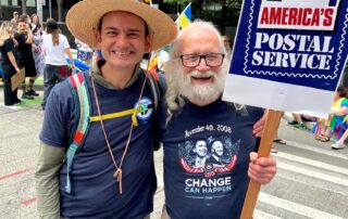 PROTEC17 member Ulysses stands with retired postal worker union sibling, Bob. They are smiling and Bob is holding a sign that reads, "SAVE AMERICA'S POSTAL SERVICE." They are standing on a crosswalk in the Pride parade.