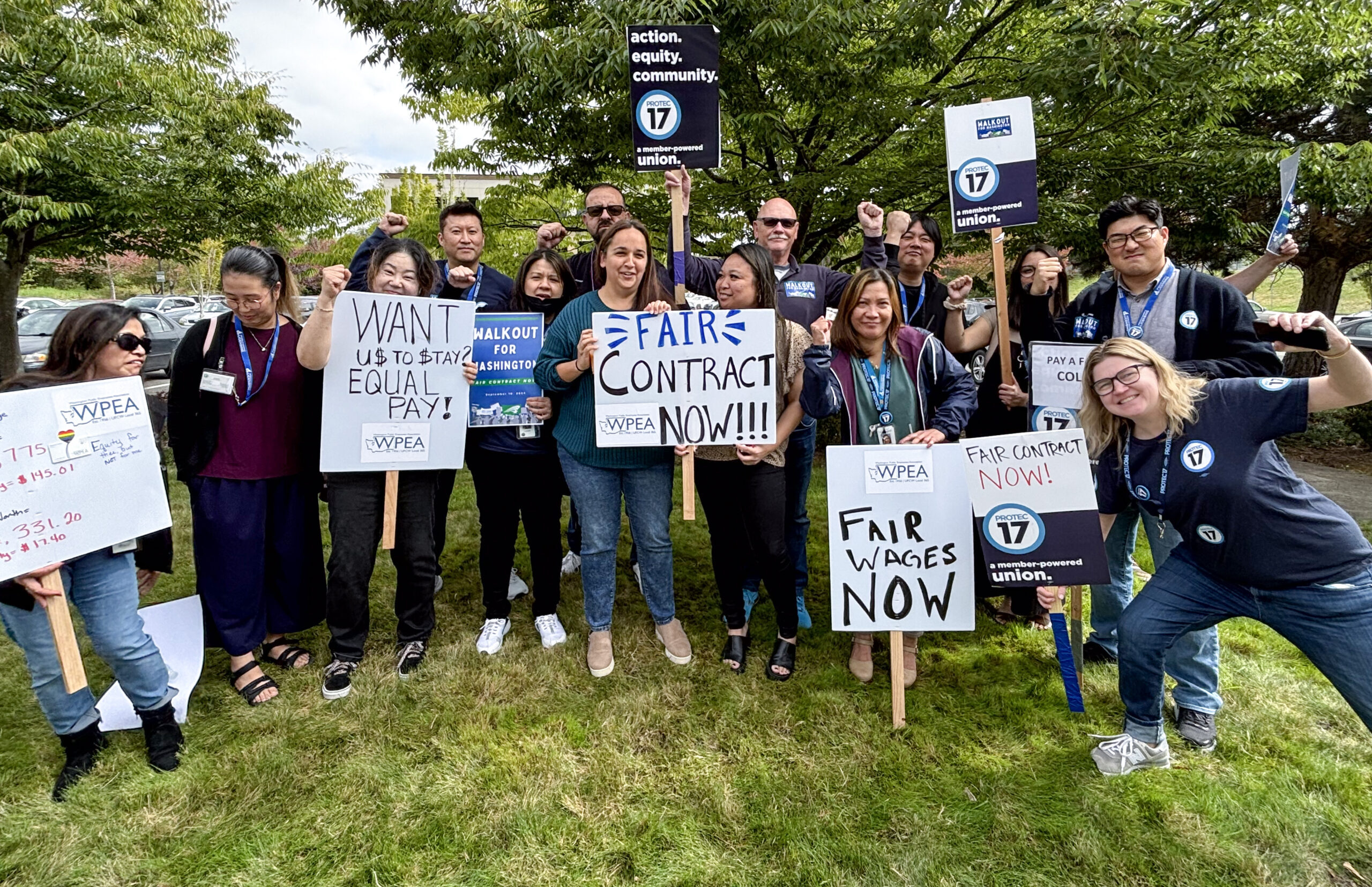 PROTEC17 members are joined by PROTEC17 staff and their WFSE and WPEA union siblings. Everyone is smiling and some folks are holding signs demanding a fair contract or raising their fists in solidarity. They are standing on the grass in front of big, green trees.