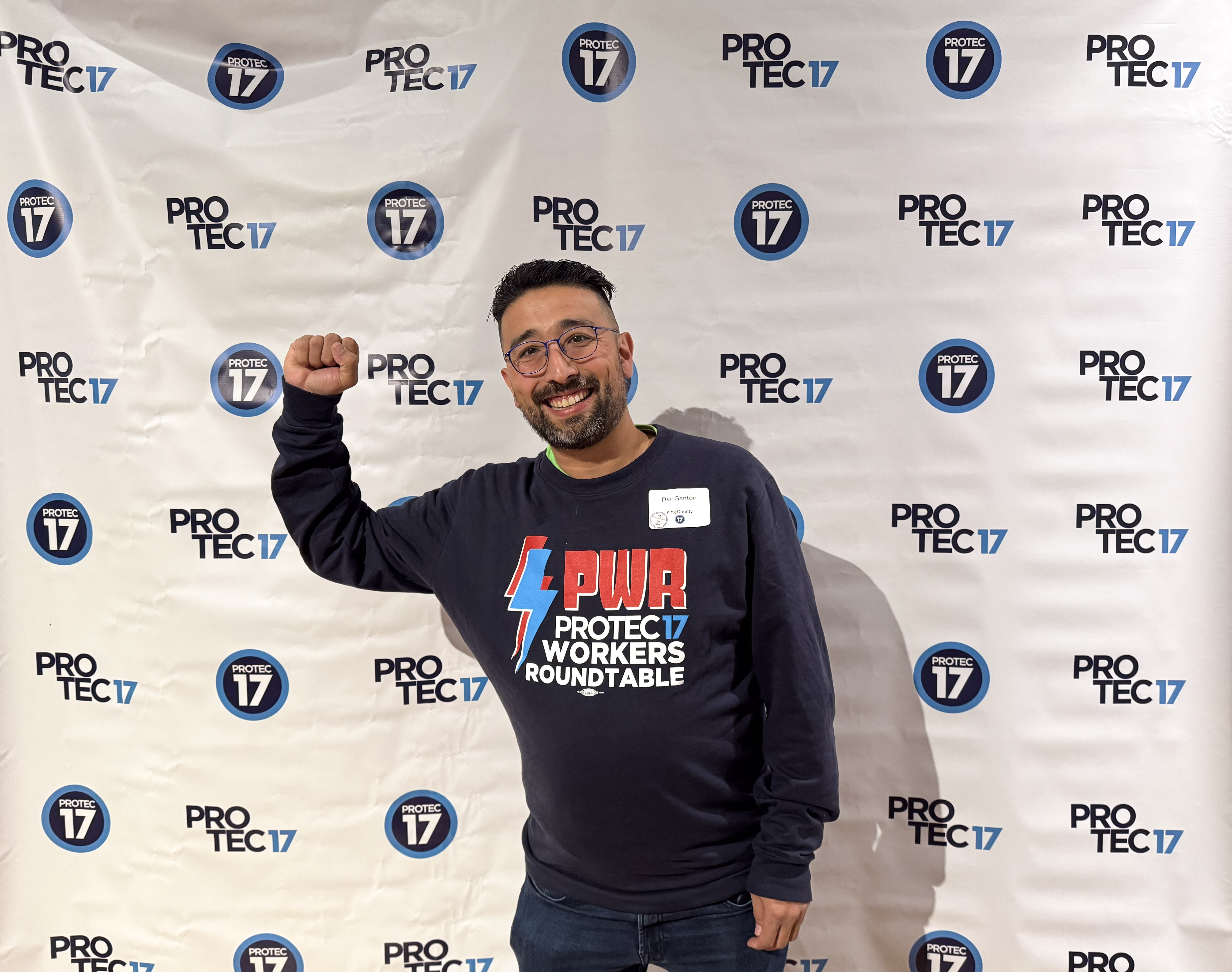 PROTEC17 member leader Dan stands in front of the PRTOEC17 backdrop that features both styles of logos. Dan is smiling and raising his fist in solidarity. He is wearing his PWR (PROTEC17 Workers Roundtable) long sleeve.