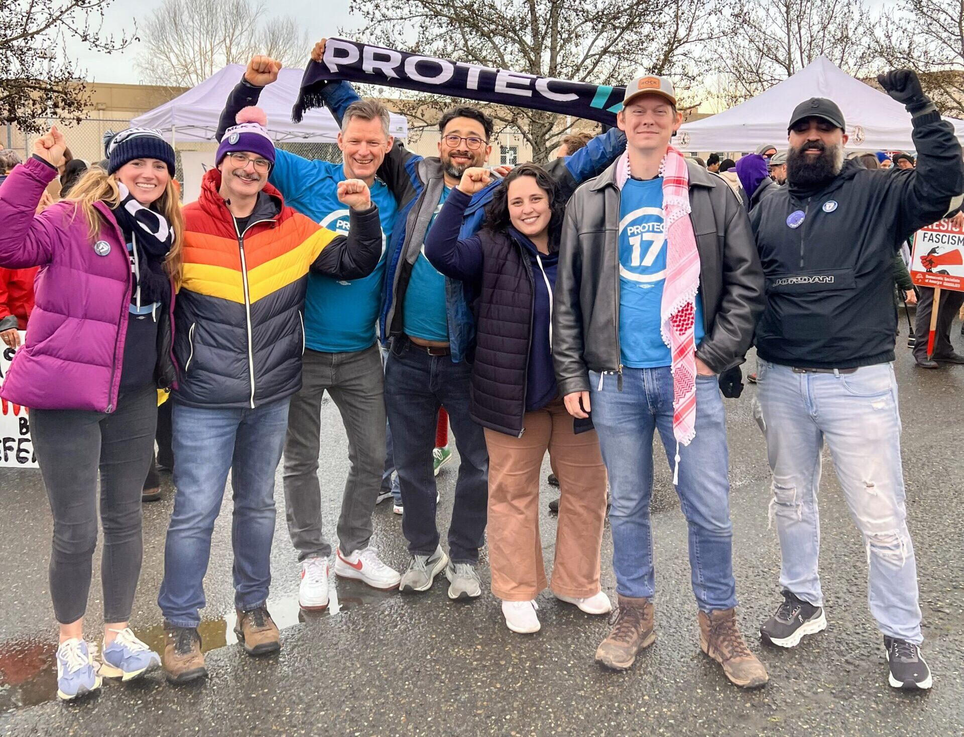 PROTEC17 members and staff stand together outside the Northwest Detention Center in Tacoma, WA. Everyone is warm clothing like jackets and/or hats, some of them PROTEC17 union gear. Everyone is smiling. One person is holding up their PROTEC17 scarf and a few folks are raising their fists in solidarity.